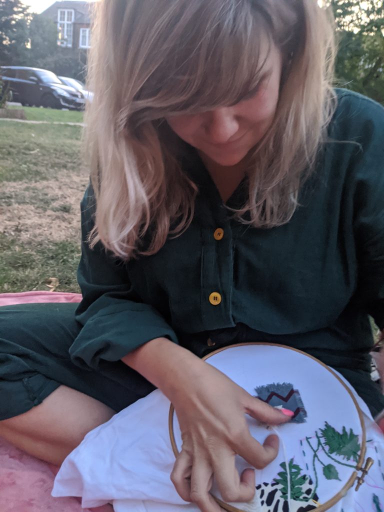 Jamie is wearing a green shirt dress and she is sat on the grass with a circular embroidery frame, she has her head bent down looking over her sewing, she long blond hair framing her face and thick fringe covering her eyes. 