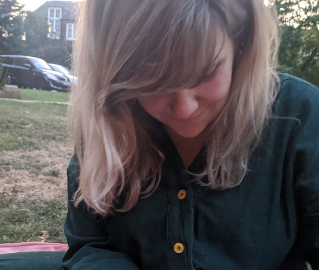 Jamie is wearing a green shirt dress and she is sat on the grass with a circular embroidery frame, she has her head bent down looking over her sewing, she long blond hair framing her face and thick fringe covering her eyes.