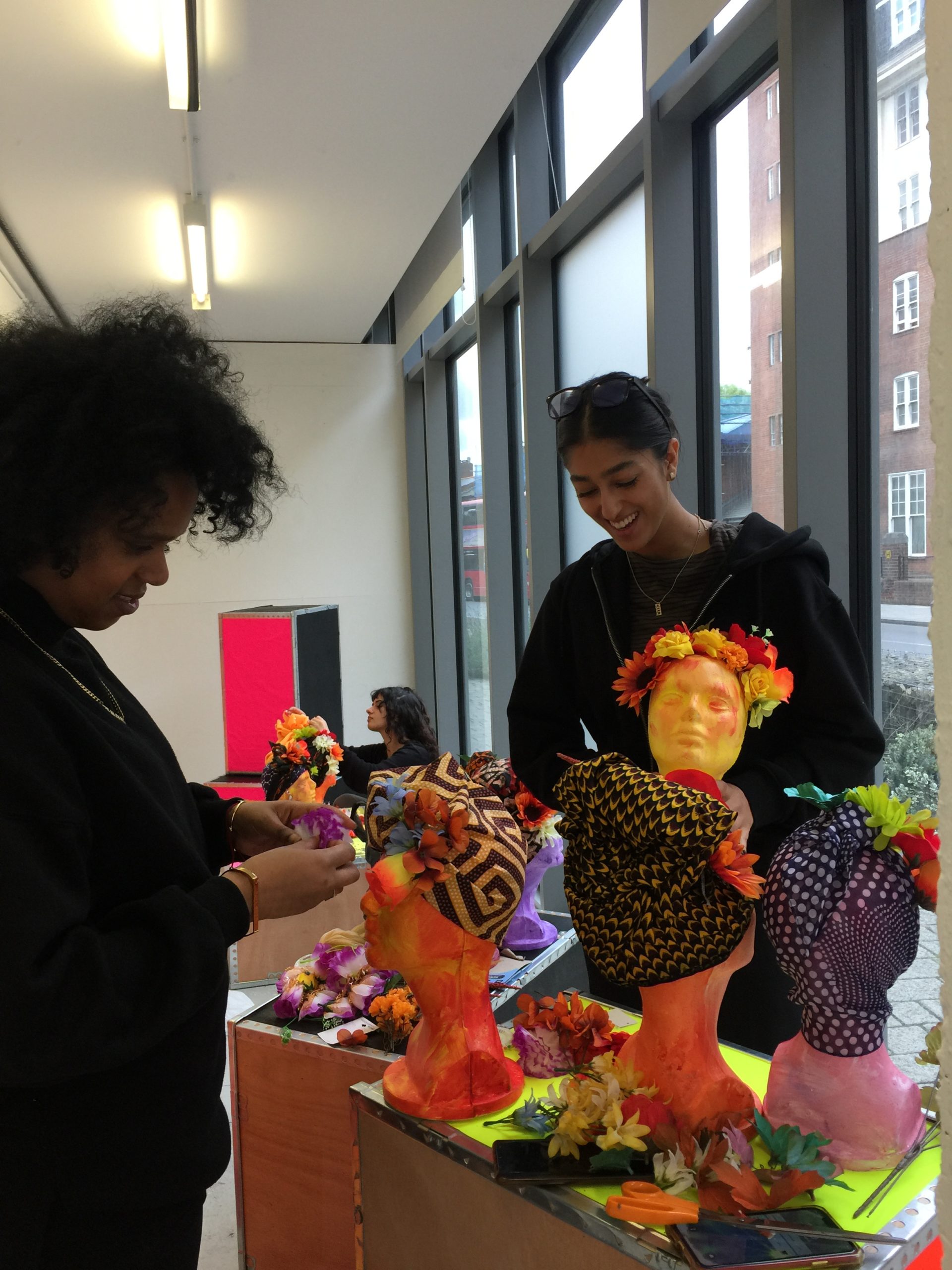 Azarra and Safa, two black women are setting up the window space in Deptford X. They are stood around large black boxes which have painted foam heads displayed on.
