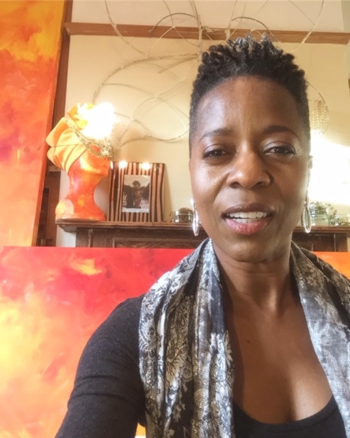 The image shows Linett, a black slim woman with short hair. She is looking directly at the camera, wearing a black scoop neck top, a black & white scarf and silver hoop earrings. In the background you can see her painting which are swirls of reds, oranges, yellows.