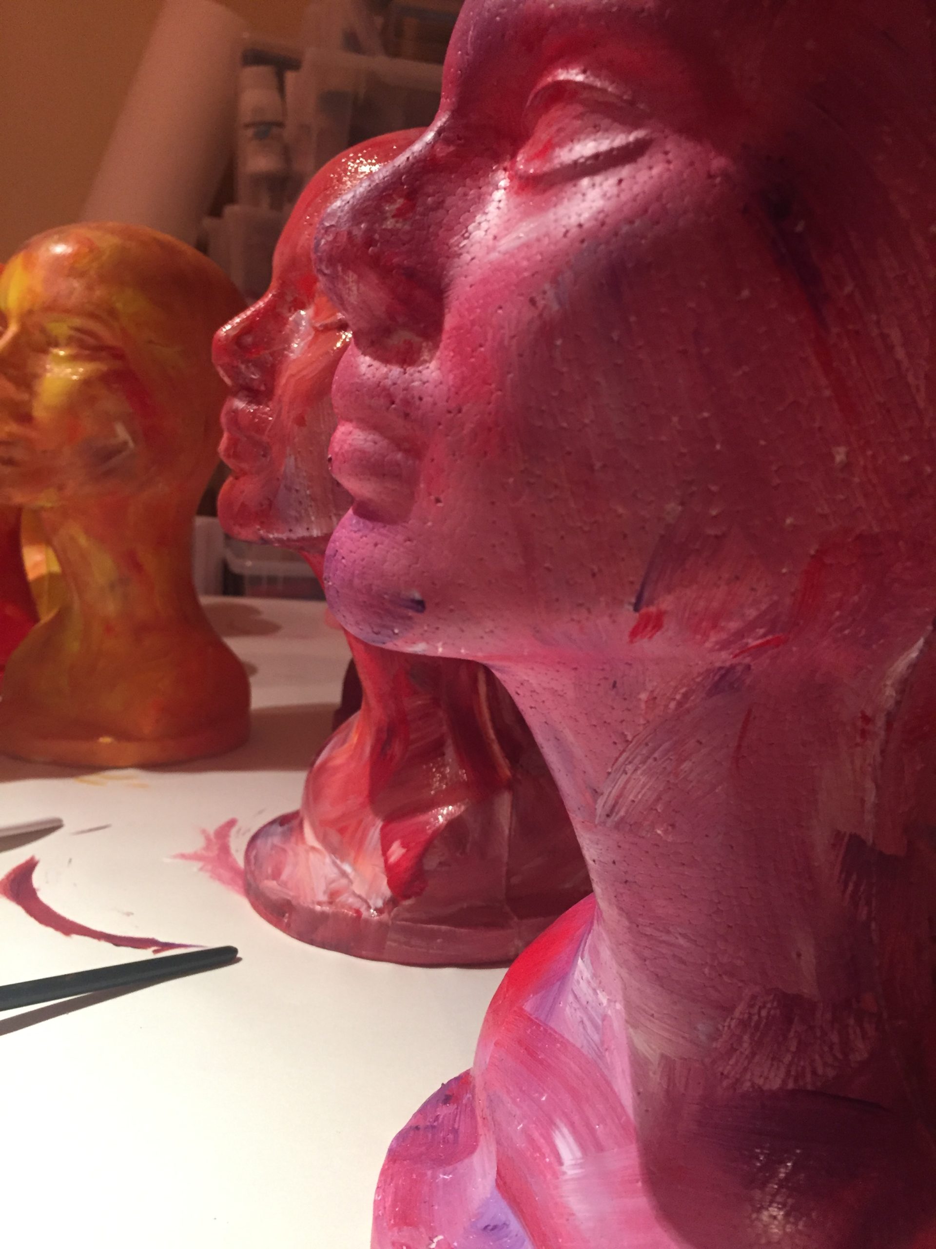 Three foam head forms in a row, each head is spray painted a different colour, pink, red, orangey gold.
