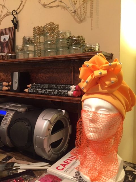 A white mannequin head sat upon a sideboard next to a large stereo, the mannequinn head is dressed in an orange headdress with an orange netted mesh over it's face.