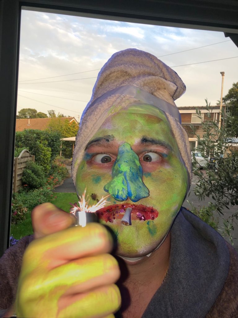 A close up image of a white male with his face crudely painted in yellow and a green painted fake and overly large and exaggerated noise with red lips, his eyes are crossed and he is holding up a lighter to light a roll up cigarette which he is holding between his lips. His hand is also smeared in the same yellow paint that is on his face, the paint is patchy on his hands. He is also sporting a beige towel on his head and appears to be wearing a dressing gown. In the background you can see blue skies with white clouds and greenery of trees and grass in the distance