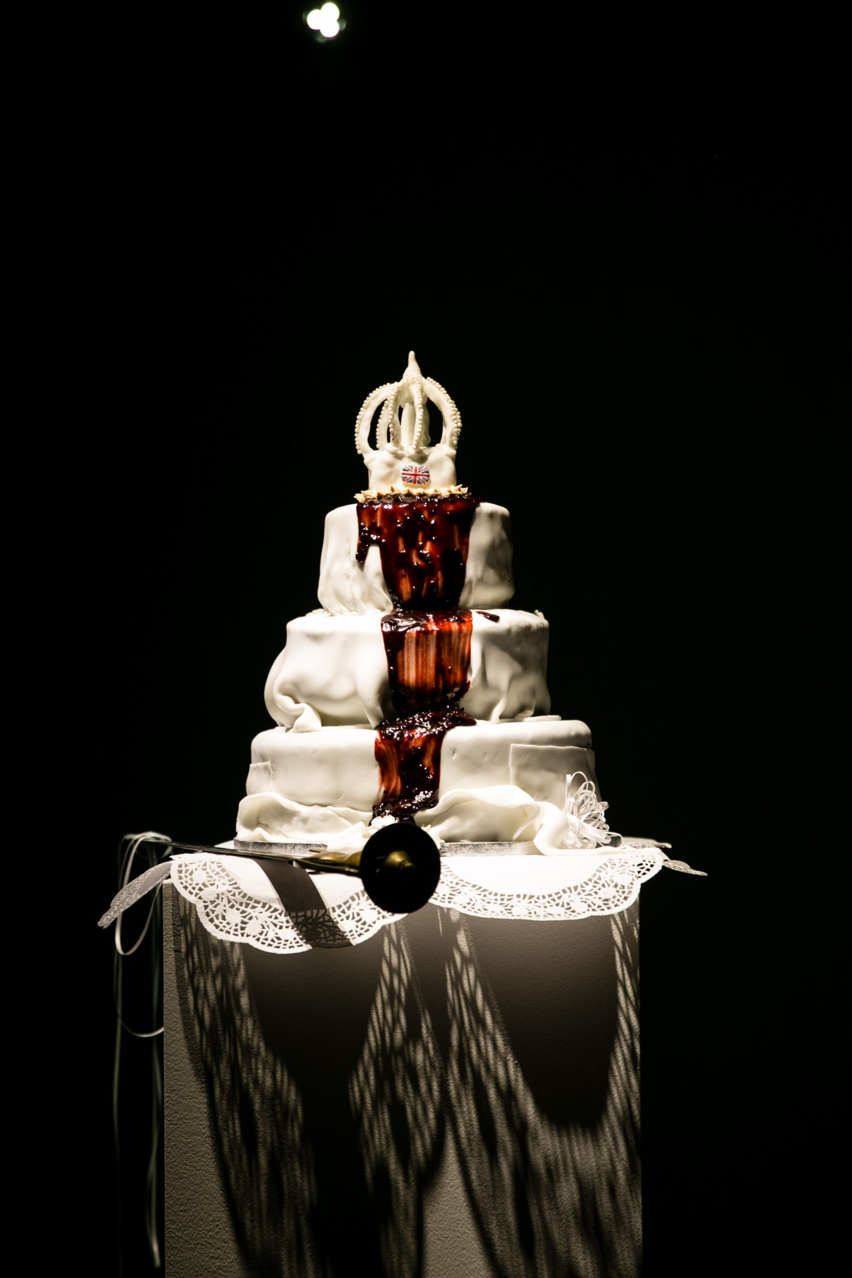 The there is black background with white table that has a three tiered cake in thick white fondant icing, with a white crown cake topper on the top tier. This cake topper is also made of the same thick icing and has a union flat painted in the center of the crown's band. Down the center of the cake, from beneath the crown, spilling down the three tiers is a thick red, blood-like substance. The cake is on a round table with a white cloth and a large white round dollie.