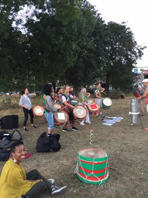Fellow Notting Hill carnivalists @baquedeaxe who have been rehearsing via webcam during lockdown, took the easing of restrictions as an opportunity to research in the park bringing music and joy in the park. There are 8 drummers, wearing large colourful drums, they are being led by a conductor with a stand-up steel drum. Linett can be seen in the bottom left-hand frame, sitting crossed legged and beaming a wide smile at the camera as the band play in a bright sunny, green park. 